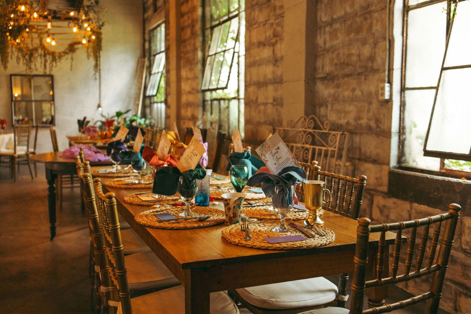 Rustic wedding reception table set with colorful napkins, gold chargers, and candlesticks in brick venue.