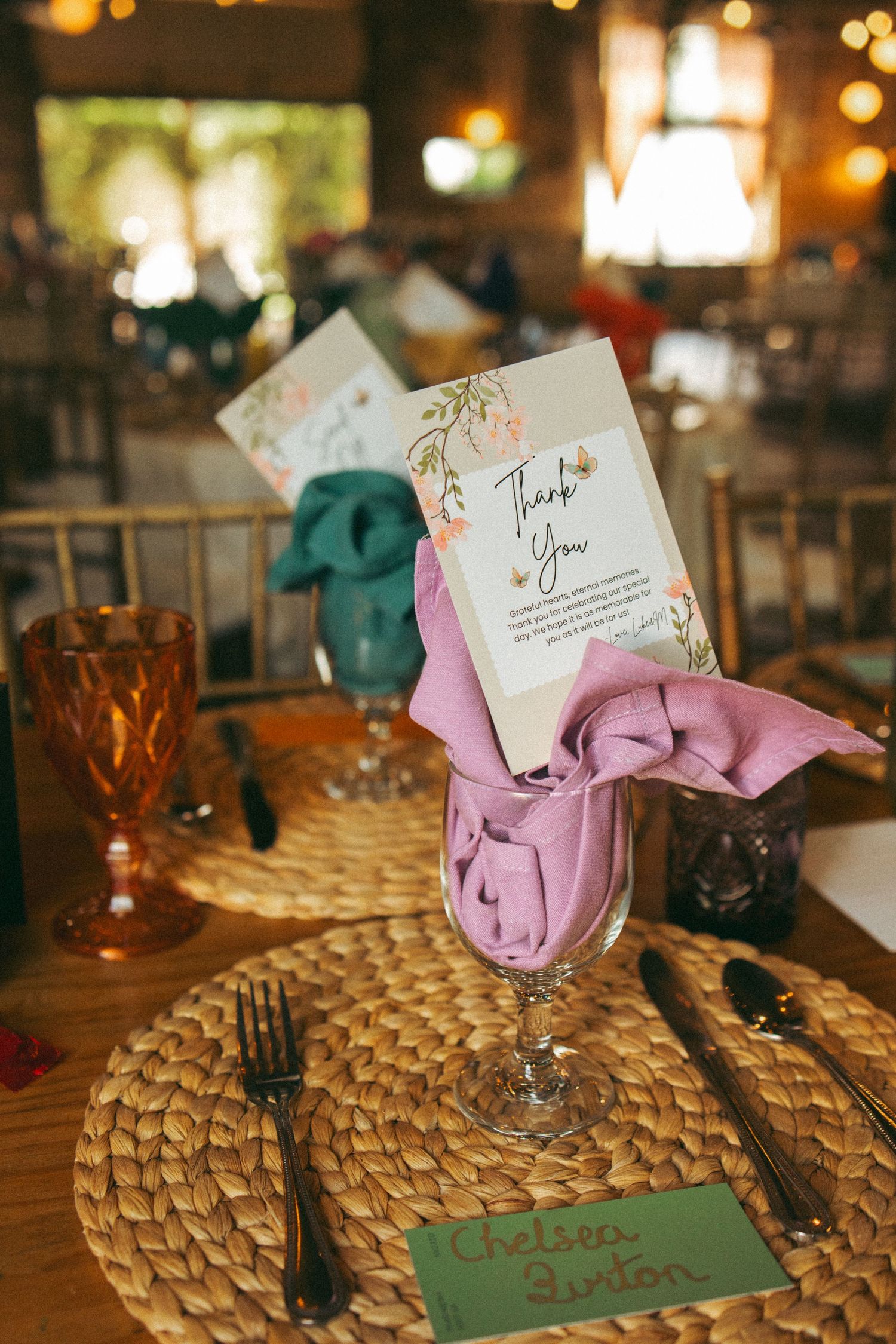 Wedding place setting with lavender napkin in wine glass, thank you card, and woven charger plate.