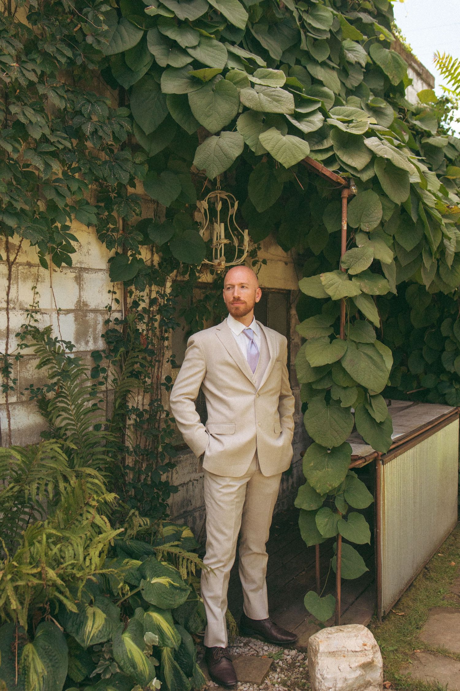 Groom in beige suit stands confidently under lush green ivy archway outdoors.