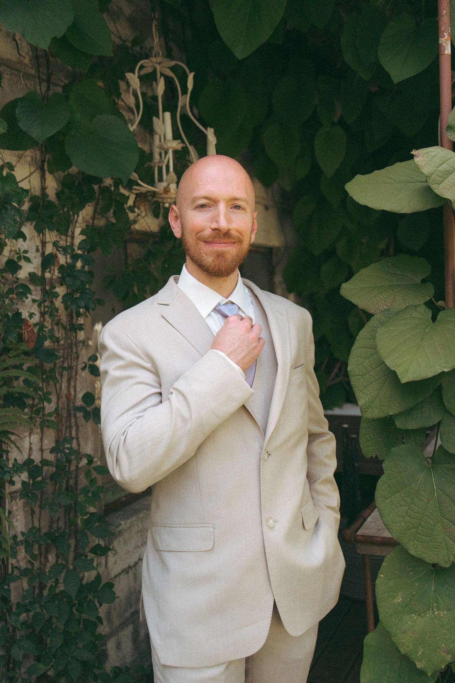 Smiling bald groom in beige suit adjusts tie surrounded by green ivy wall.