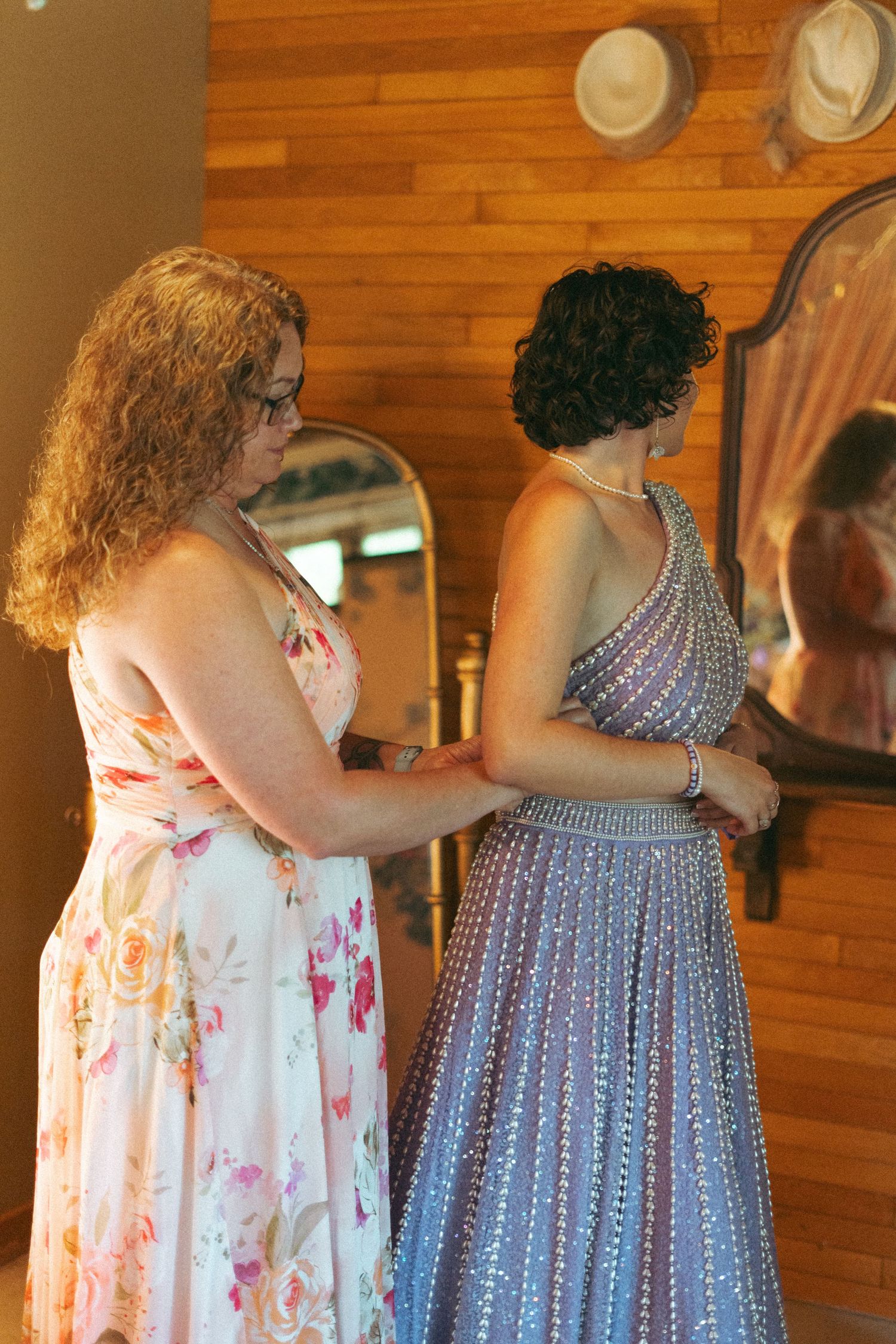 Woman in floral dress helping bride in beaded silver gown in a wood-paneled dressing room.