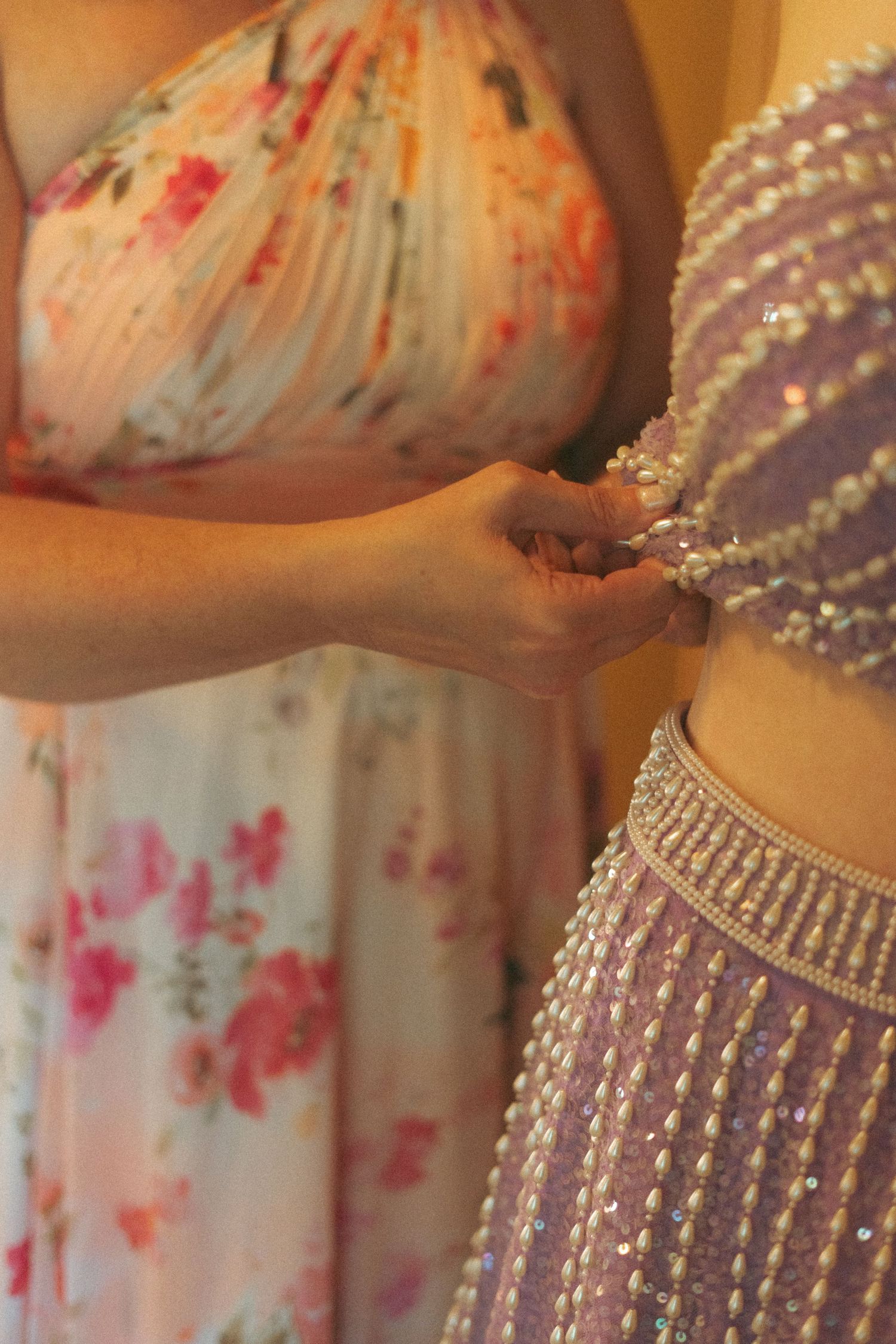 Woman in floral dress fastening buttons on a lavender pearl-embellished bridal gown.