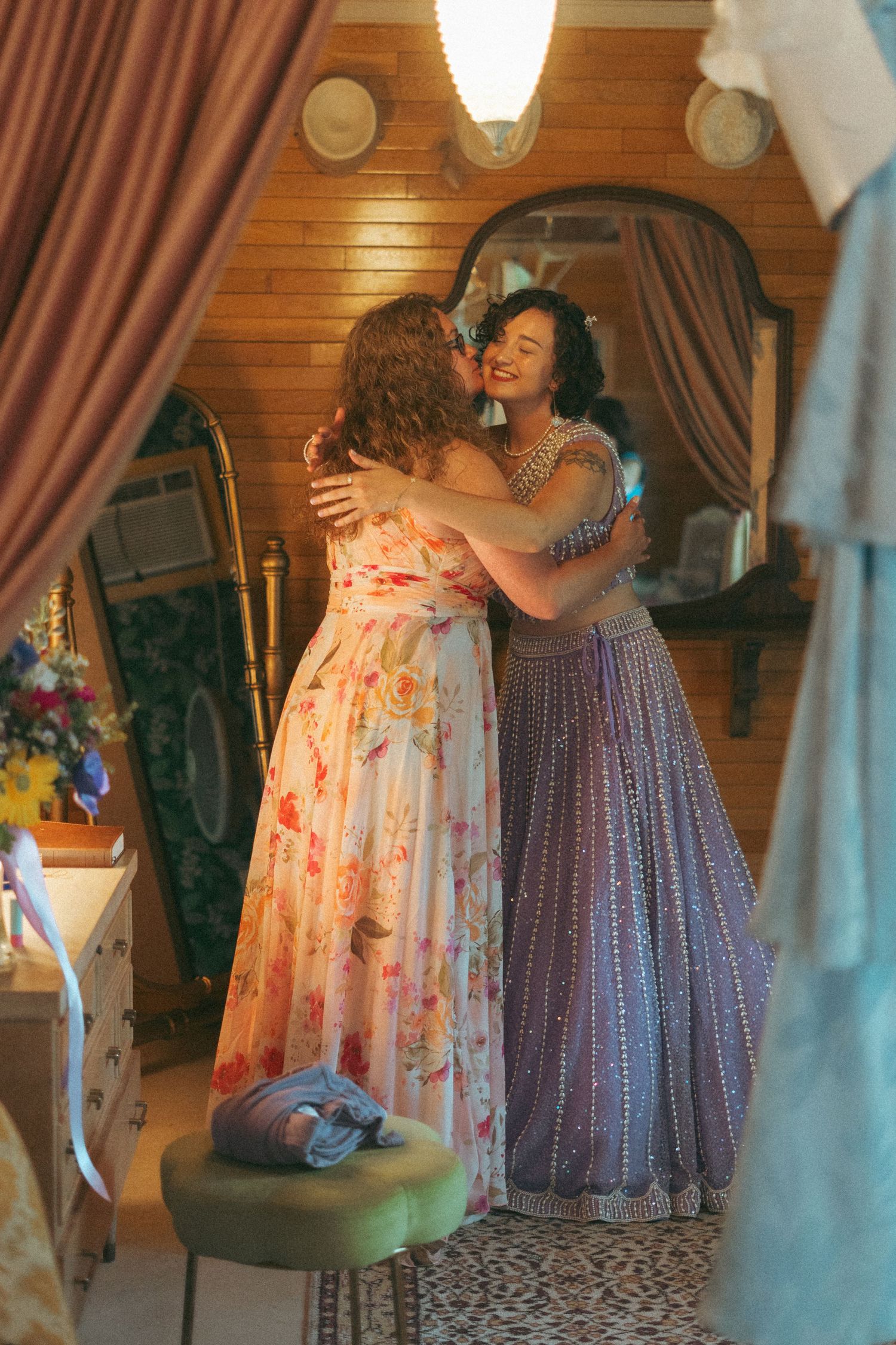 Two women in elegant gowns sharing a warm embrace in a vintage dressing room.