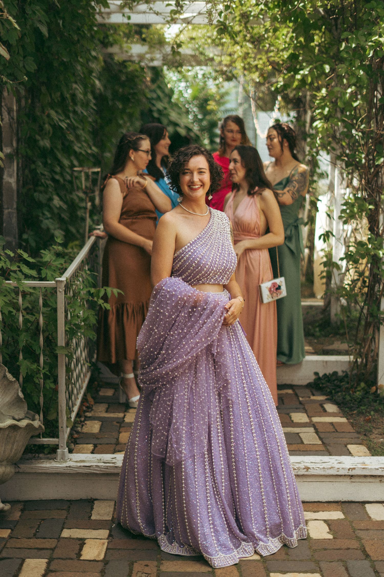 Smiling bride in lavender pearl lehenga poses with bridesmaids in colorful dresses in garden setting.