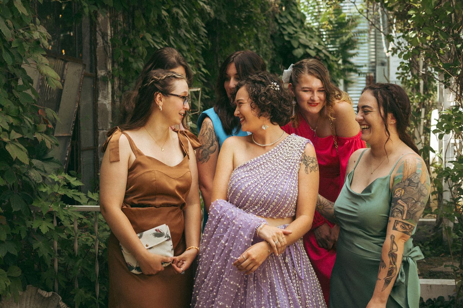 Bride in lavender lehenga laughing with four bridesmaids in colorful dresses in an outdoor garden.