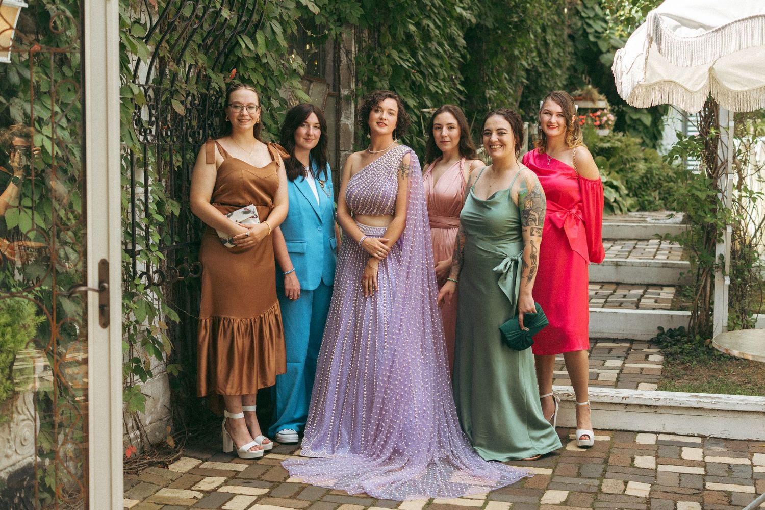 Bride in lavender beaded lehenga posing with six bridesmaids in colorful dresses in garden setting.