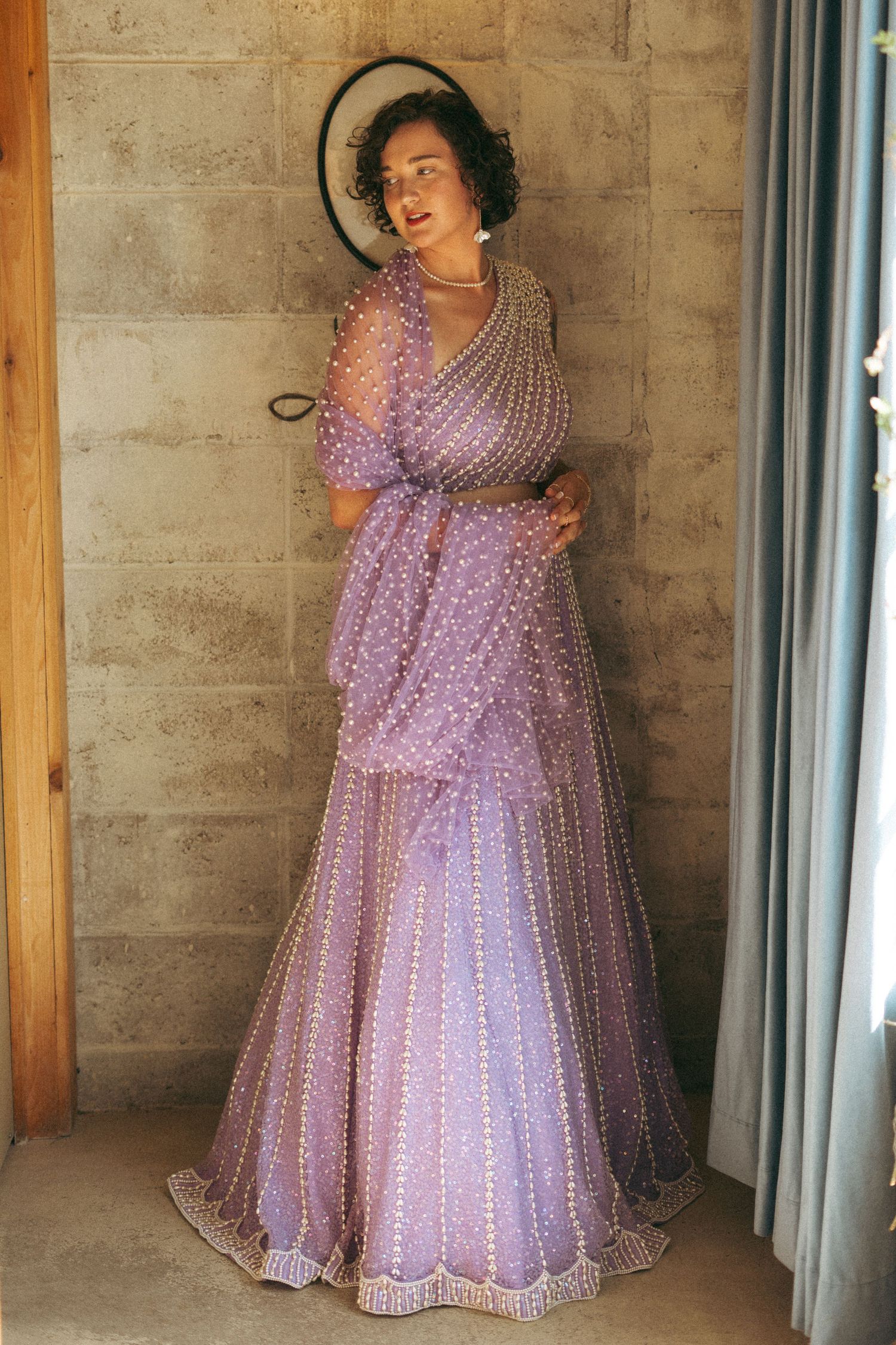 Bride in lavender pearl-embellished lehenga holding a hat and posing against a stone wall indoors.