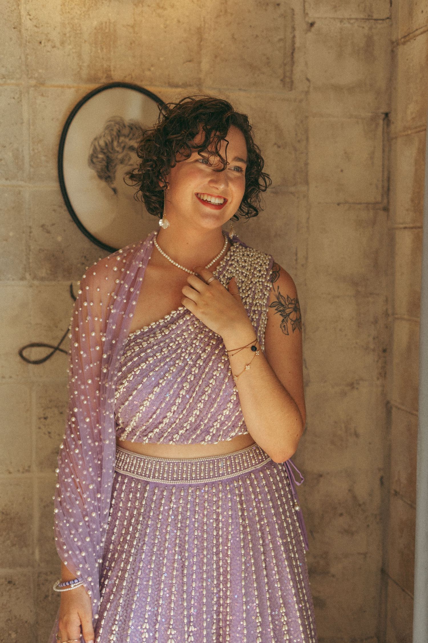 Smiling bride in lavender beaded lehenga with pearl necklace posing against a stone wall outdoors.