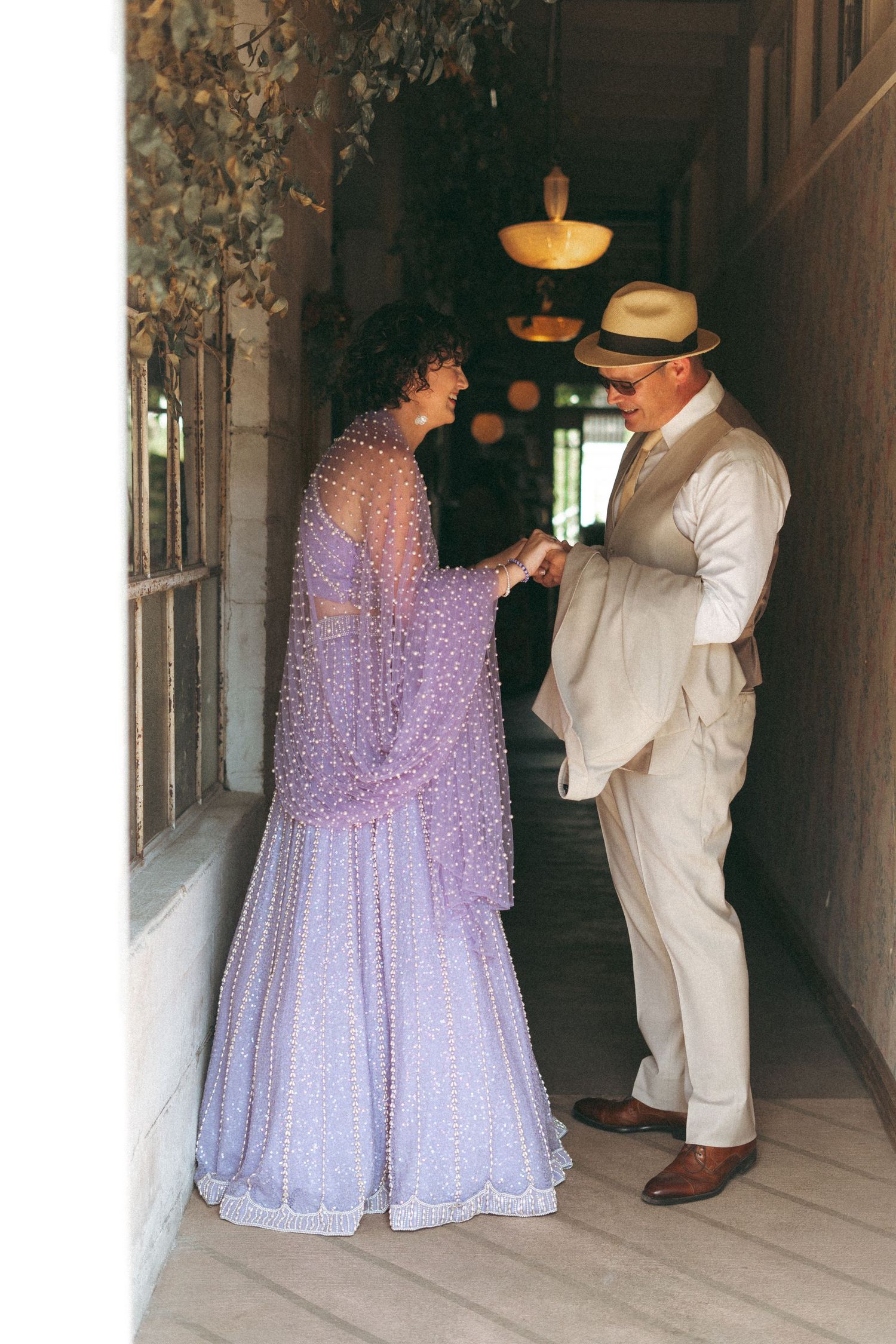 Woman in lavender beaded gown holds hands with man in beige suit and hat in elegant hallway.