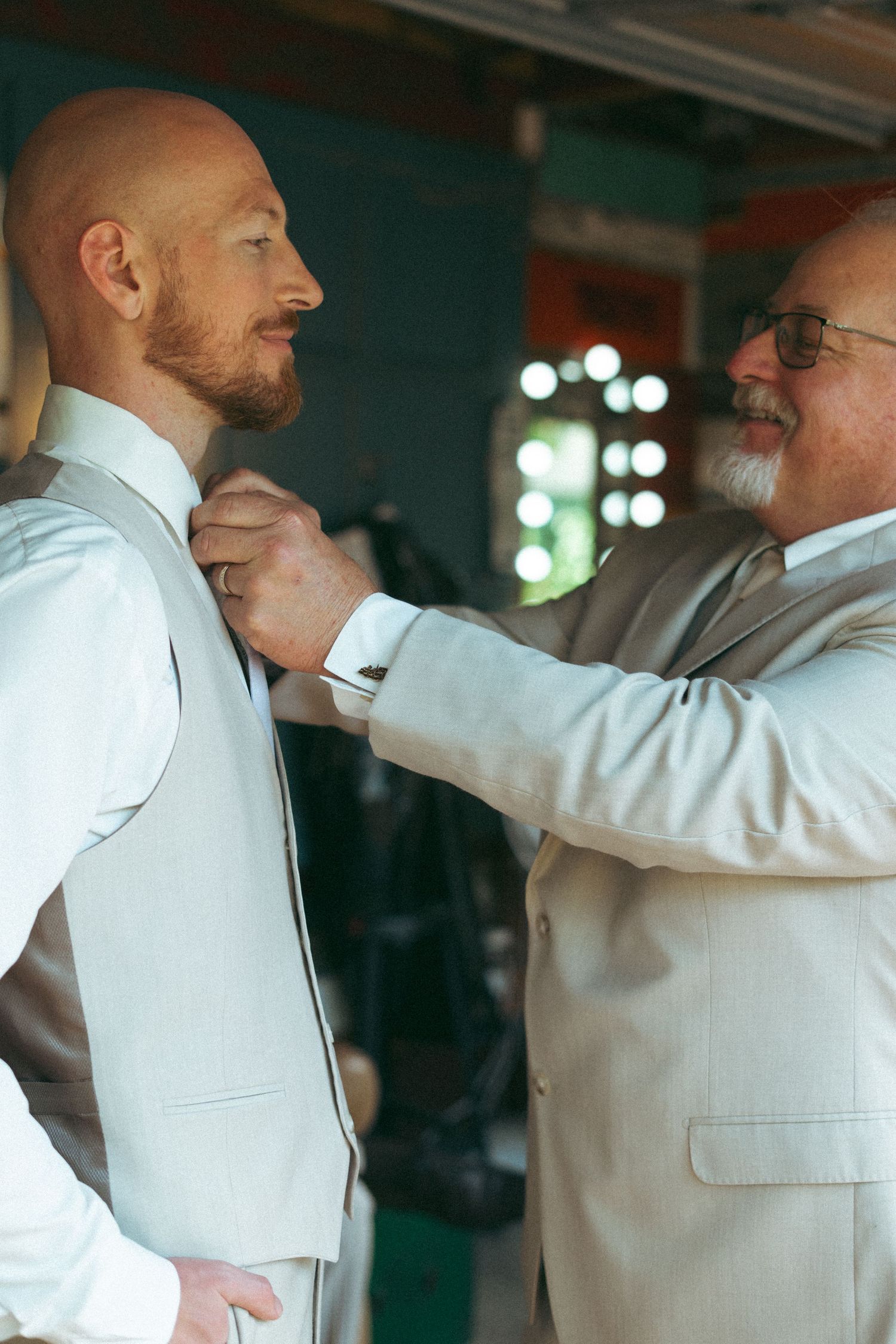 Older man in beige suit adjusts groom's collar in wedding preparation scene.