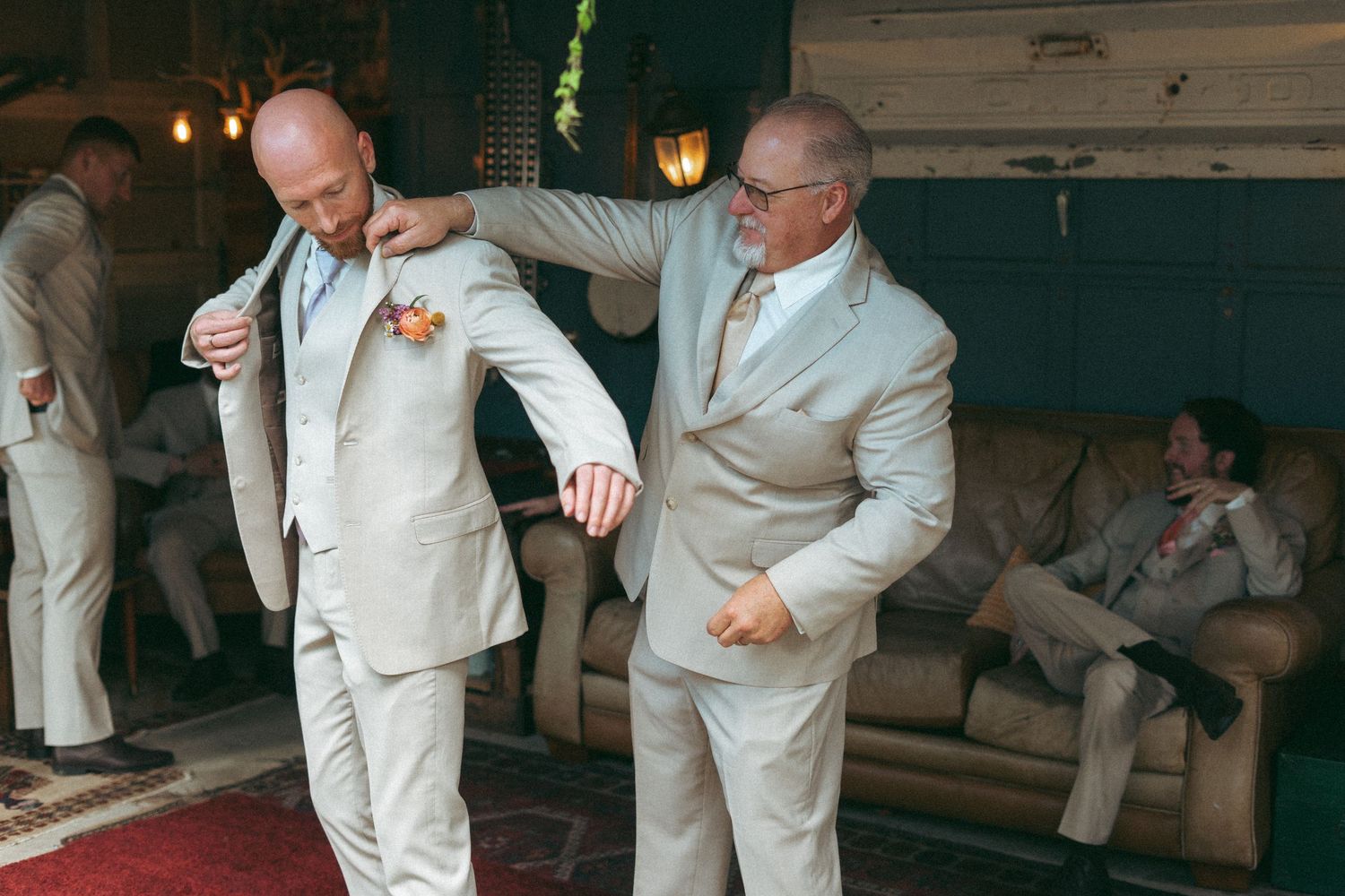 Older man helps bald groom put on beige wedding suit jacket in rustic venue.