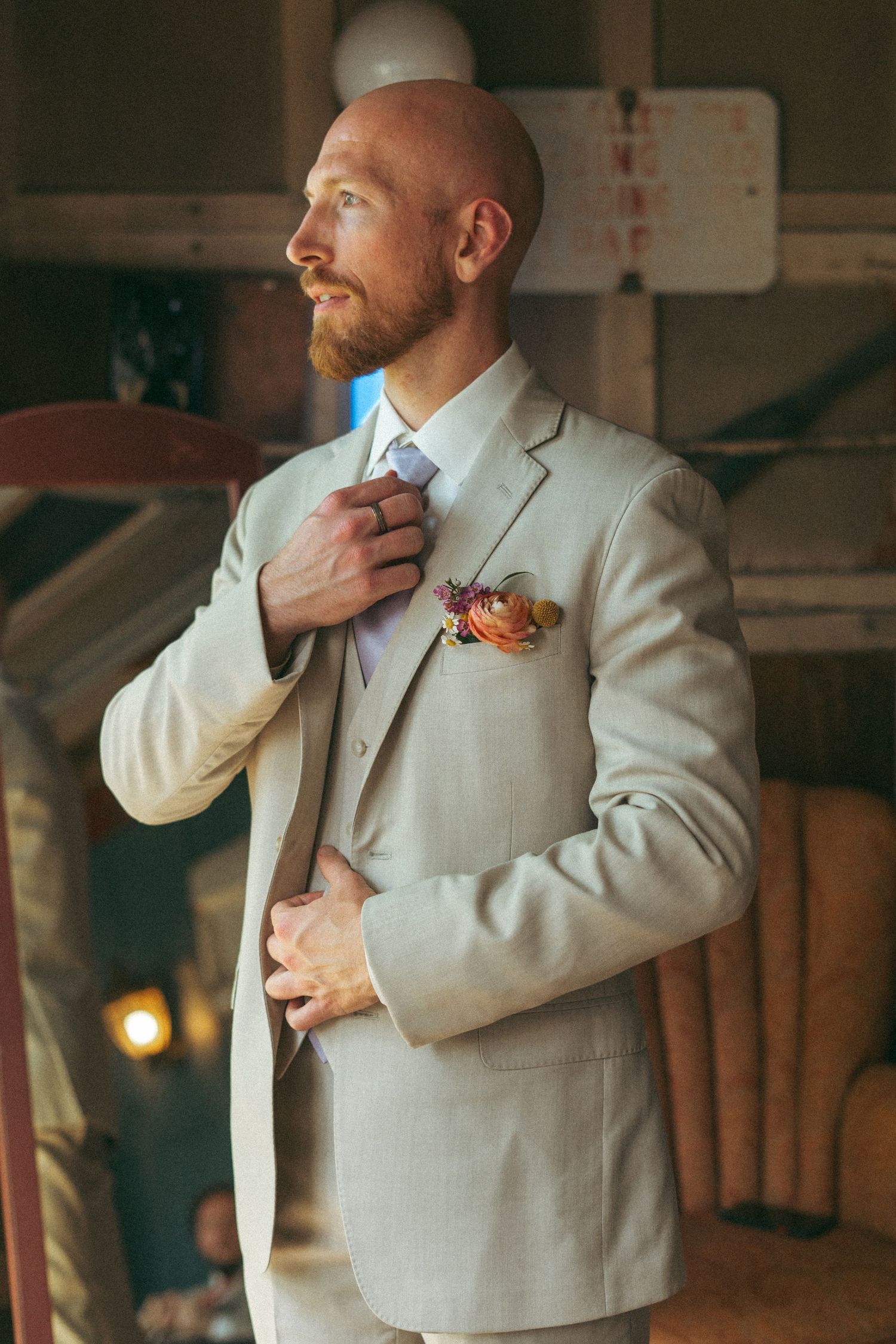Bald groom with red beard adjusts tie wearing beige suit with floral boutonniere.