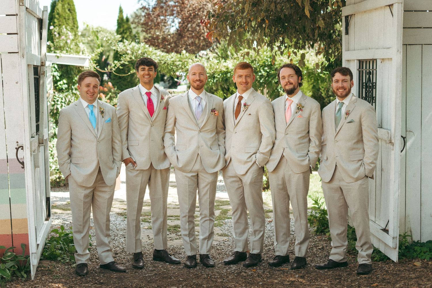 Groom and five groomsmen pose in matching beige suits with colorful ties outdoors.