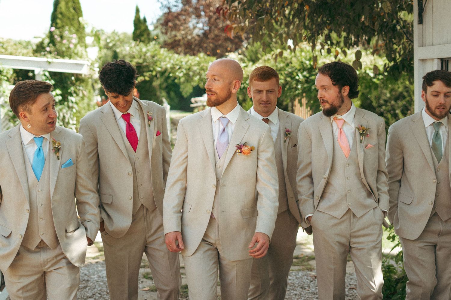 Groom and groomsmen in beige suits walk together laughing in outdoor wedding setting.