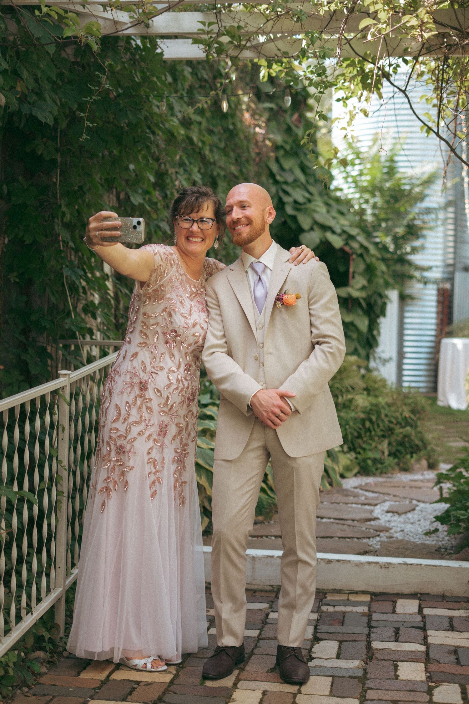Mother and bald groom take selfie together in garden, both dressed for wedding.