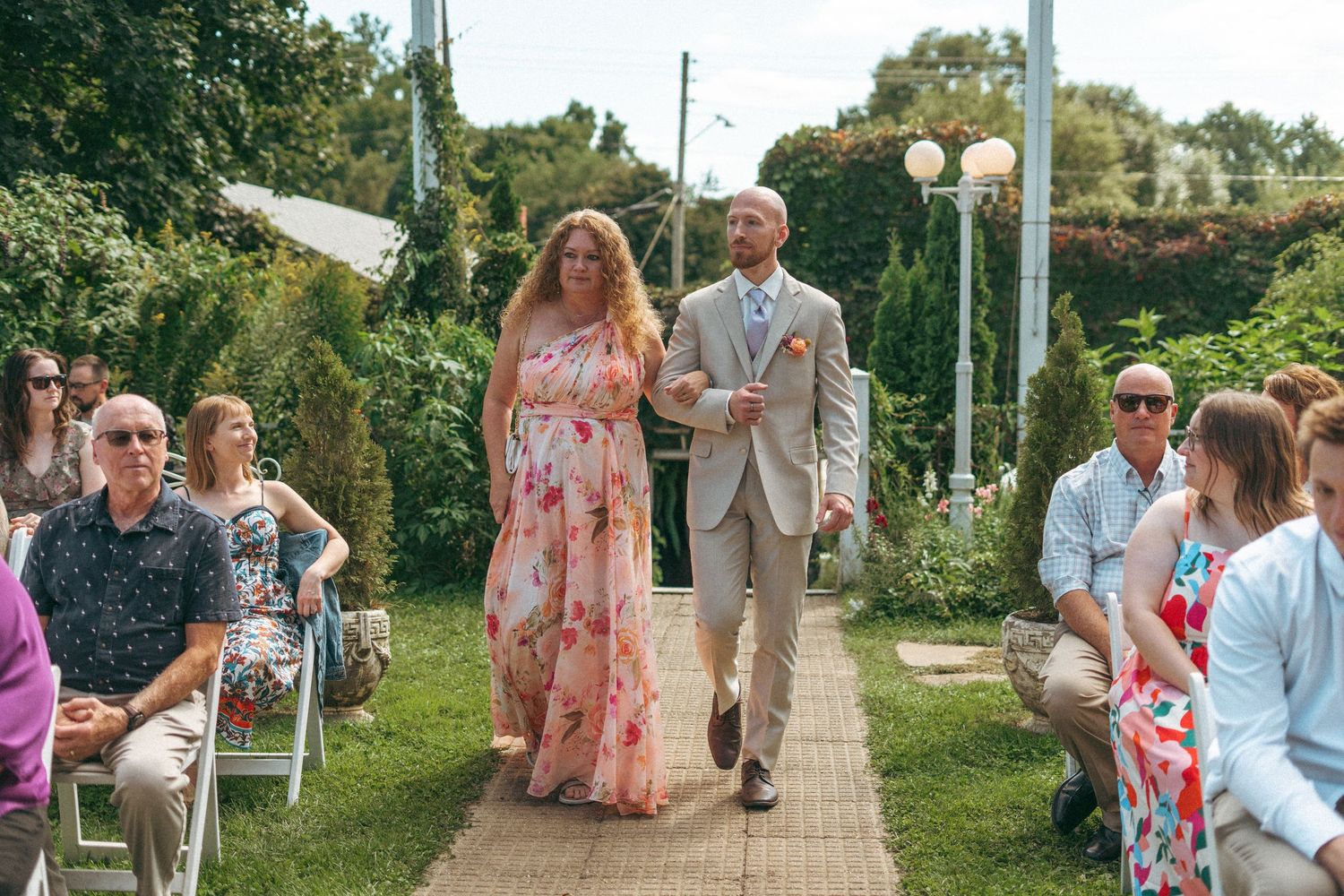 Woman in floral pink dress and man in tan suit walking down the aisle at an outdoor garden wedding.