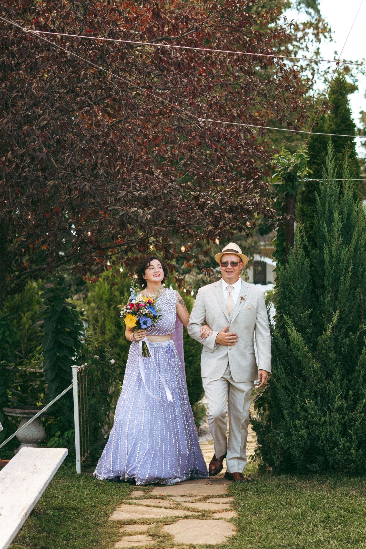 Bride in lavender sequined sari holding colorful bouquet walks down garden aisle with escort in tan suit.