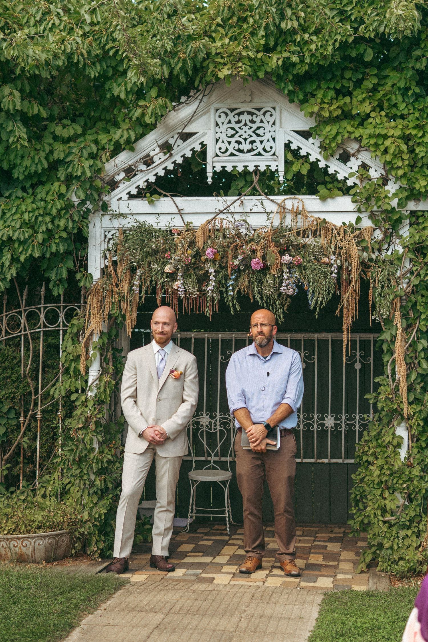 Groom in beige suit standing with officiant beneath a floral-adorned ivy-covered garden gate altar.