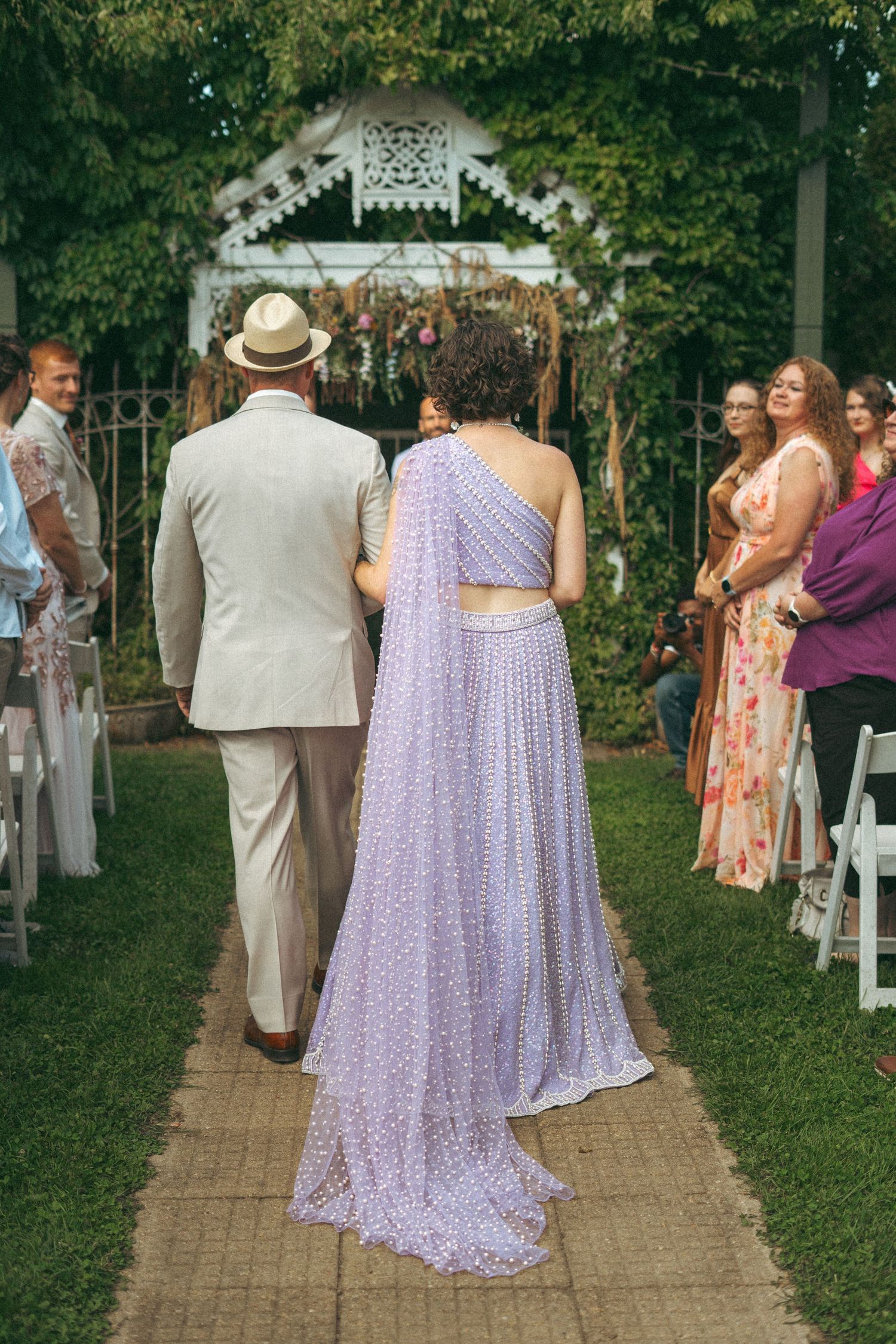 Bride in sparkling lavender sari walking down brick aisle with escort toward ivy-covered floral altar.