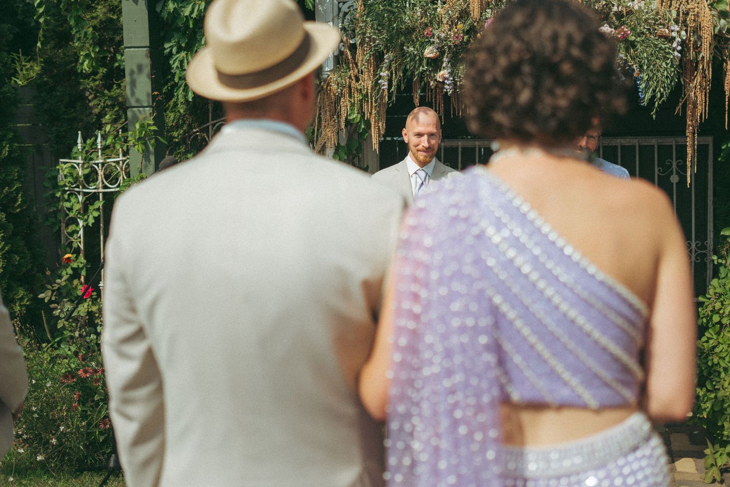 Groom smiling as bride in lavender sari approaches the altar during an outdoor garden wedding ceremony.