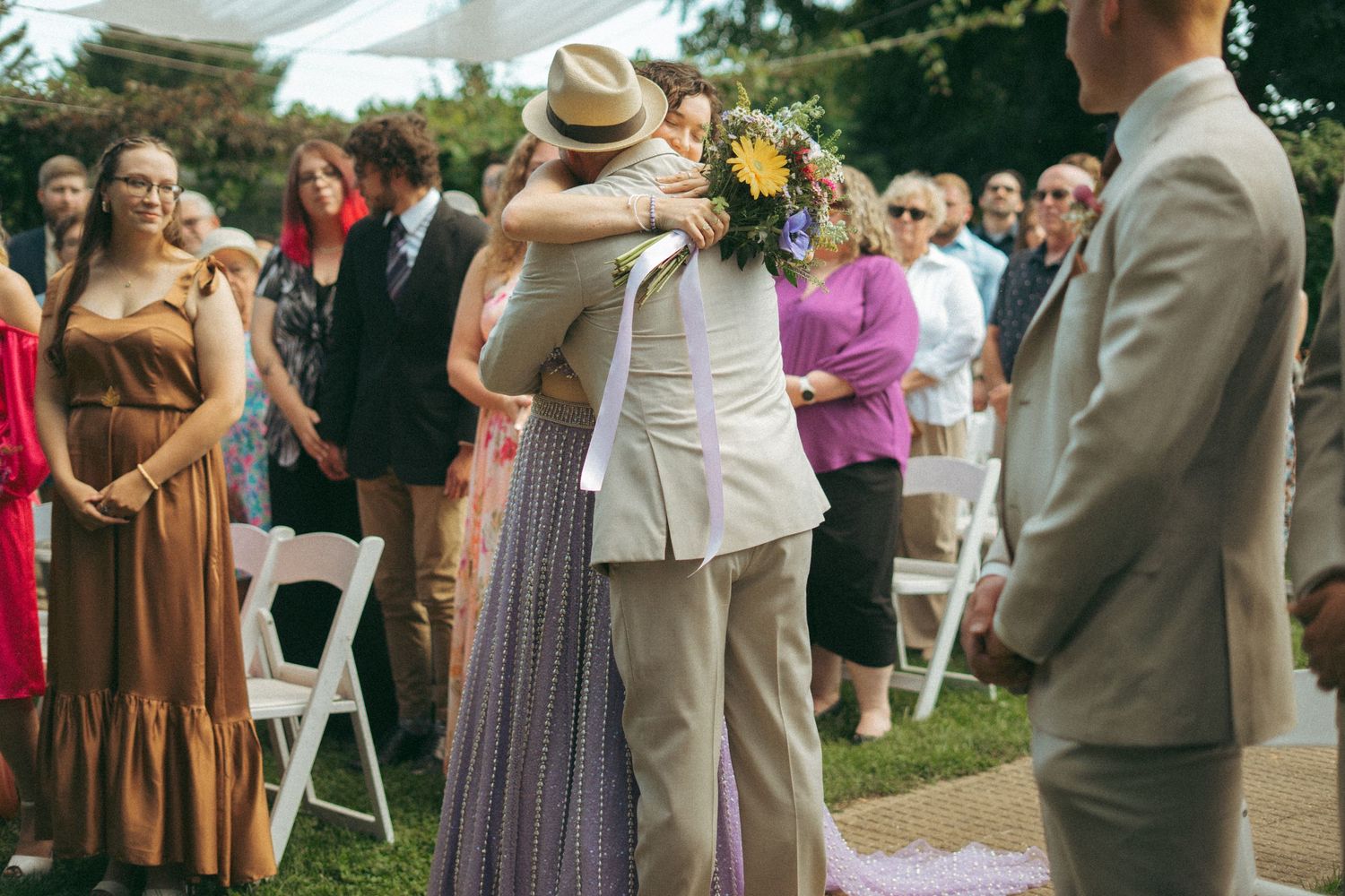 Bride in lavender gown embracing escort at the altar while groom watches at outdoor garden wedding.