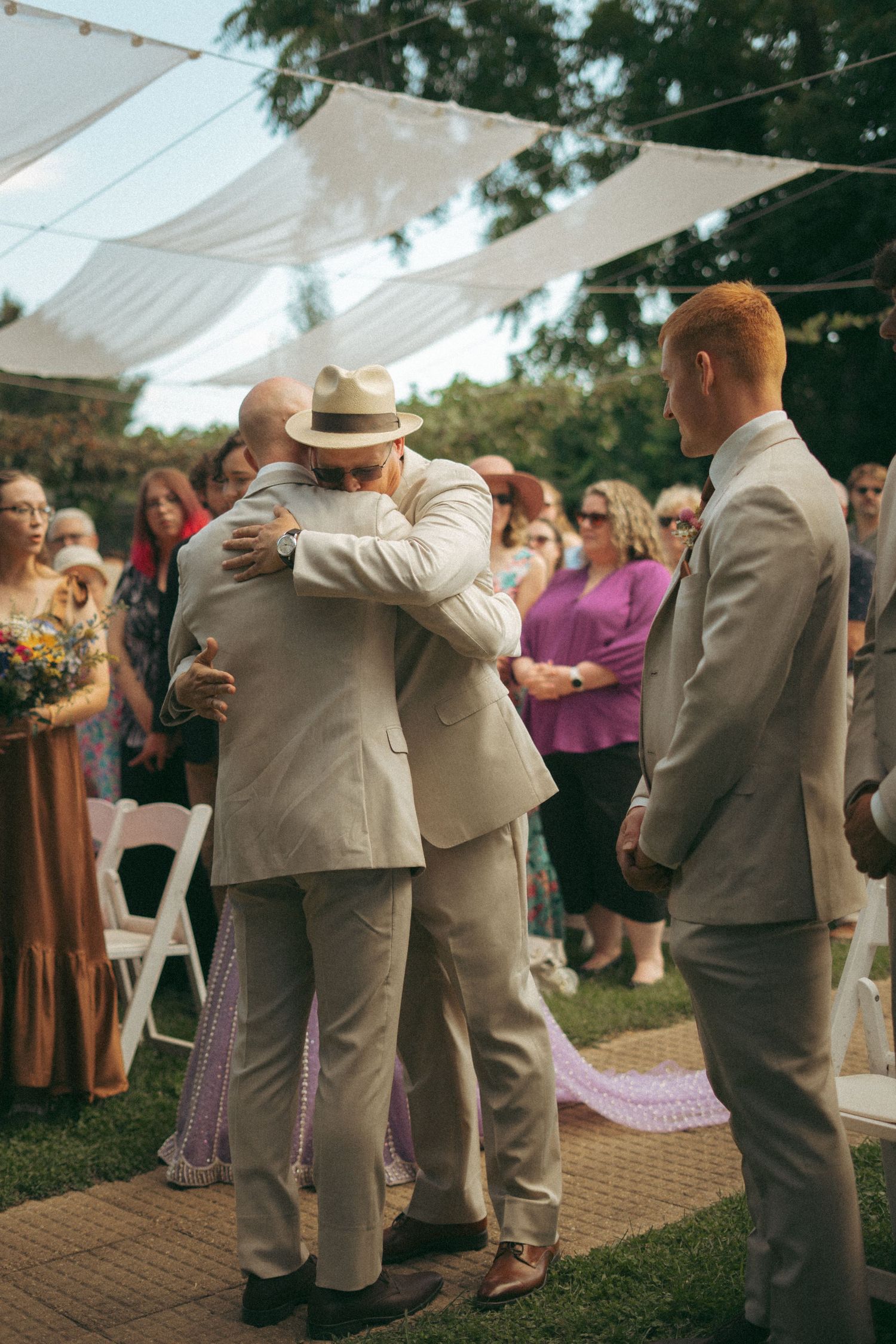Two grooms share an emotional hug during an outdoor wedding ceremony while guests look on.