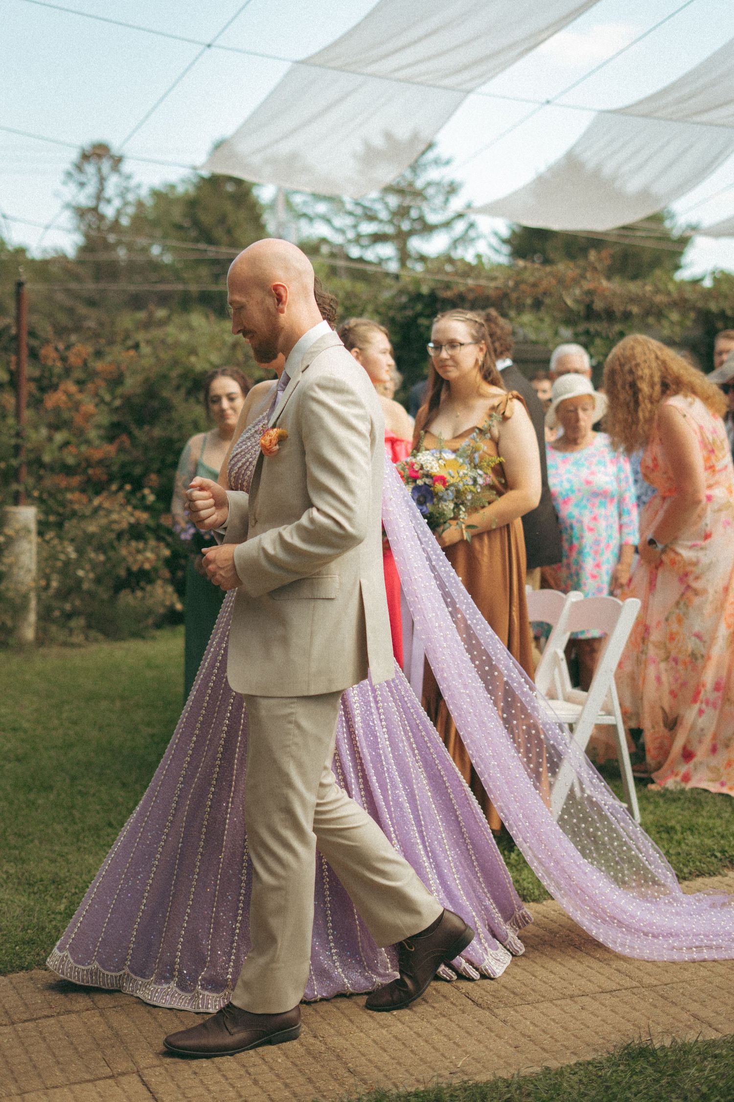 Groom escorts bride in lavender gown down the aisle at an outdoor garden wedding ceremony.