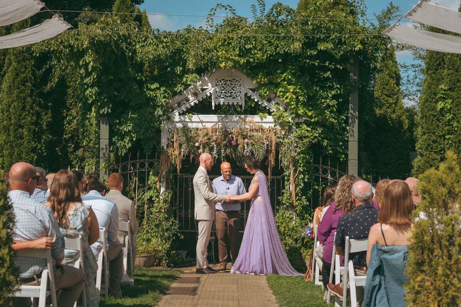 Guests seated in white chairs witness an outdoor garden wedding ceremony under a floral arch.