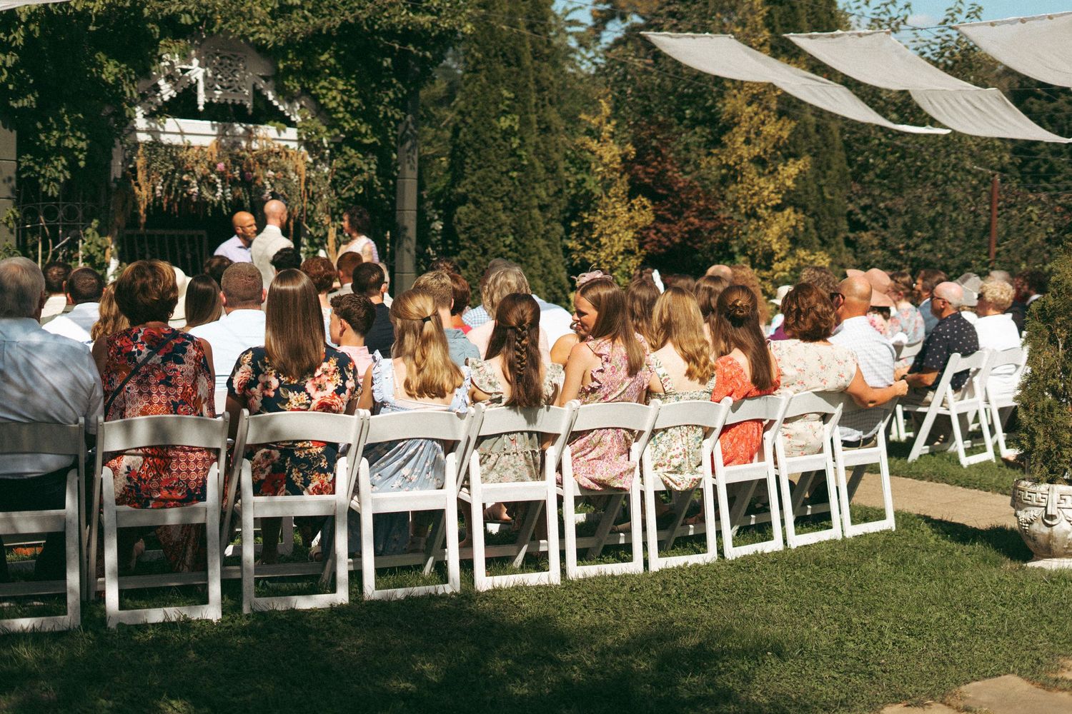 Wedding guests seated in white chairs watching an outdoor garden ceremony on a sunny day.