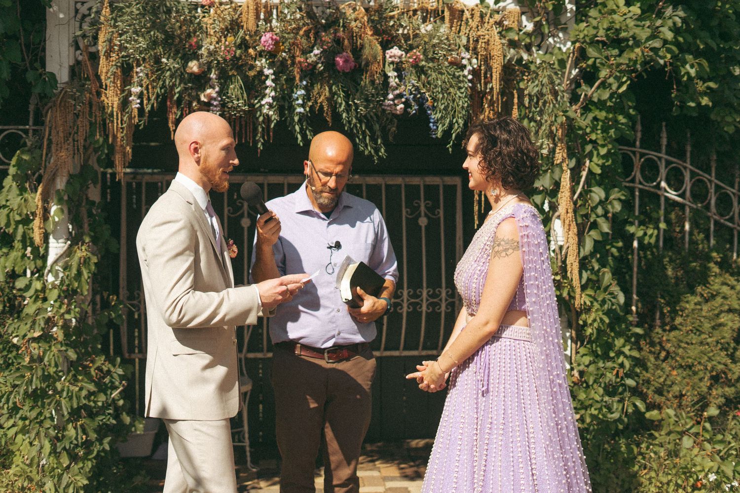 Groom reads vows to bride in lavender dress as officiant stands beside them outdoors.