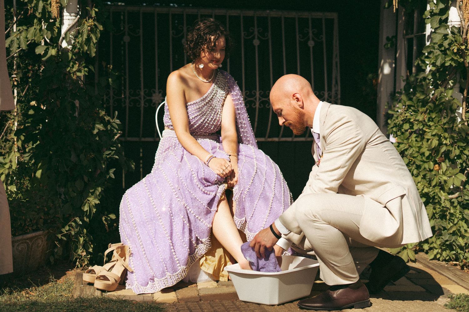Groom washing bride's feet in a basin outdoors, bride wearing lavender Indian bridal gown at garden wedding.