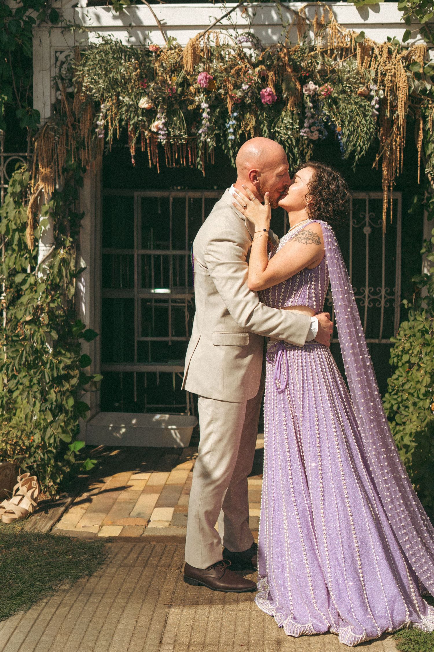 Bride and groom sharing first kiss under floral arch, bride in lavender lehenga, groom in beige suit.