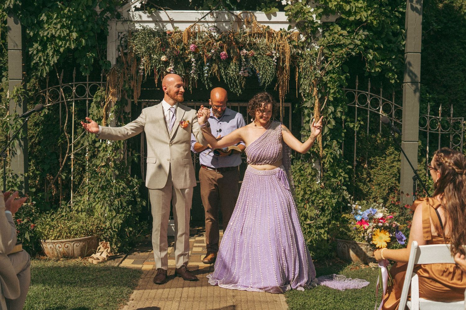 Newlyweds celebrating with officiant under flower-adorned pergola at outdoor garden wedding ceremony.