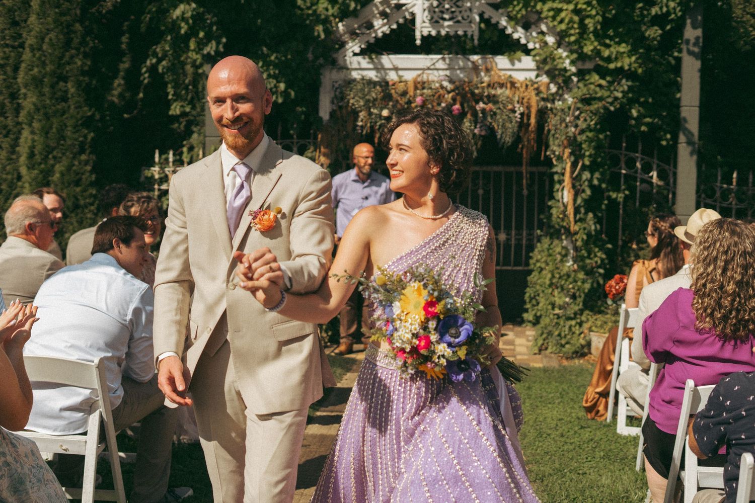 Bride in lavender lehenga and groom in tan suit walking down aisle holding colorful bridal bouquet.