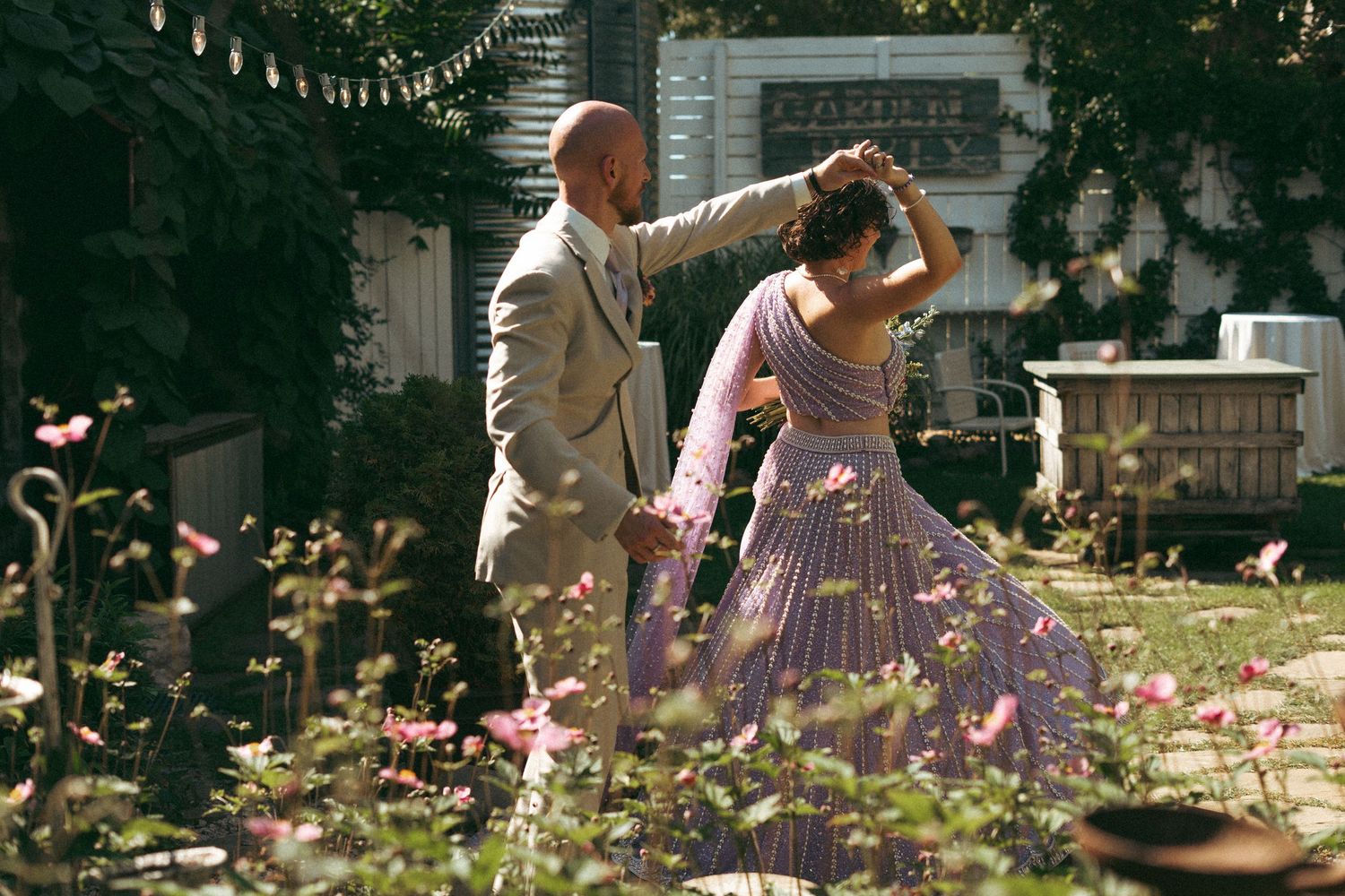 Groom twirling bride in lavender embroidered lehenga among blooming flowers in romantic garden setting.