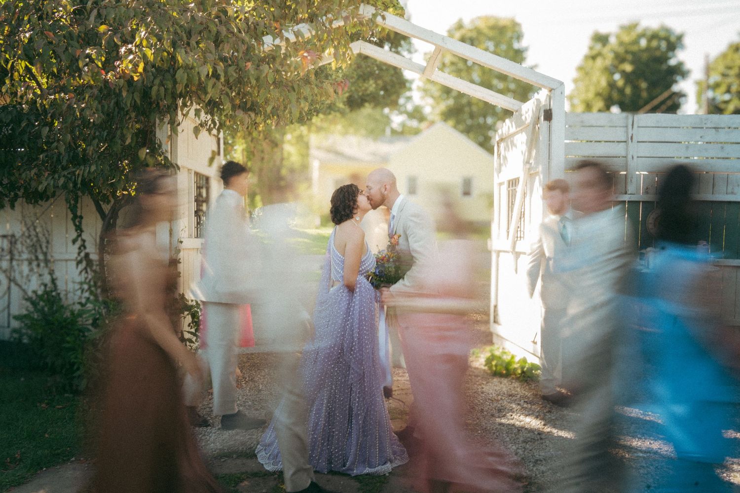Artistic motion-blur photo of bride and groom kissing surrounded by moving wedding guests outdoors.