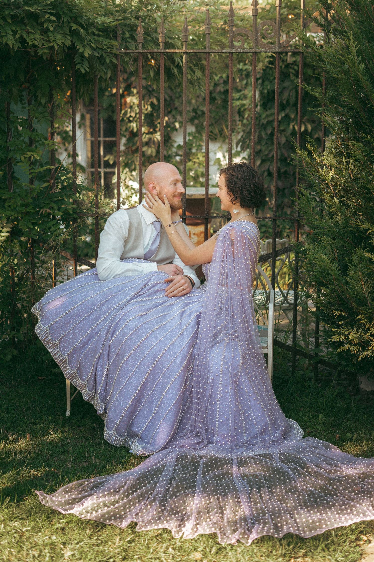 Bride in lavender embroidered lehenga sitting with groom on bench surrounded by lush green garden foliage.