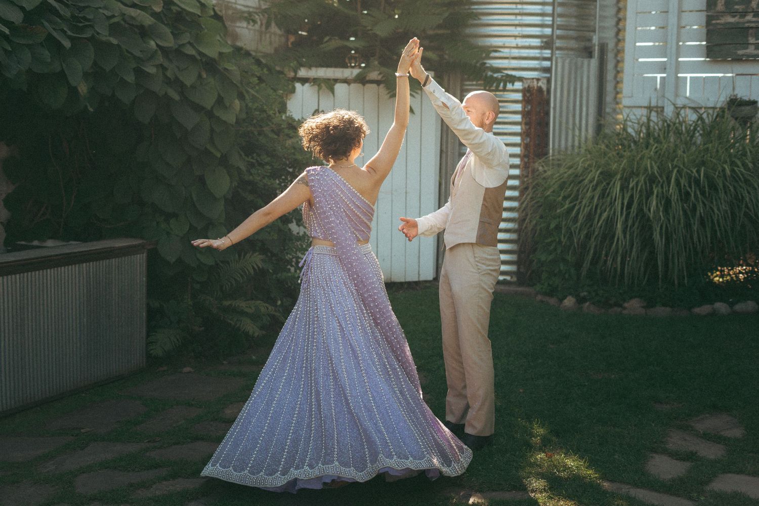 Bride in lavender lehenga and groom in beige suit dancing together in sunlit backyard garden setting.
