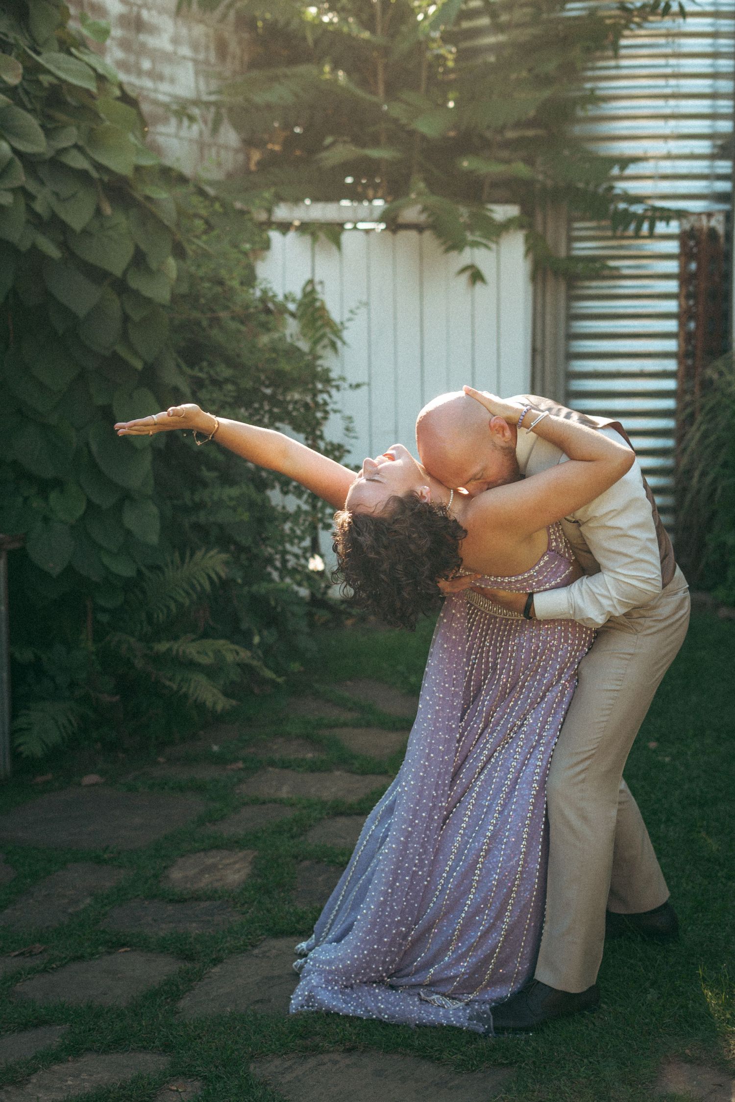 Couple sharing a romantic dip kiss in a lush garden, bride in lavender beaded gown extending her arm gracefully.