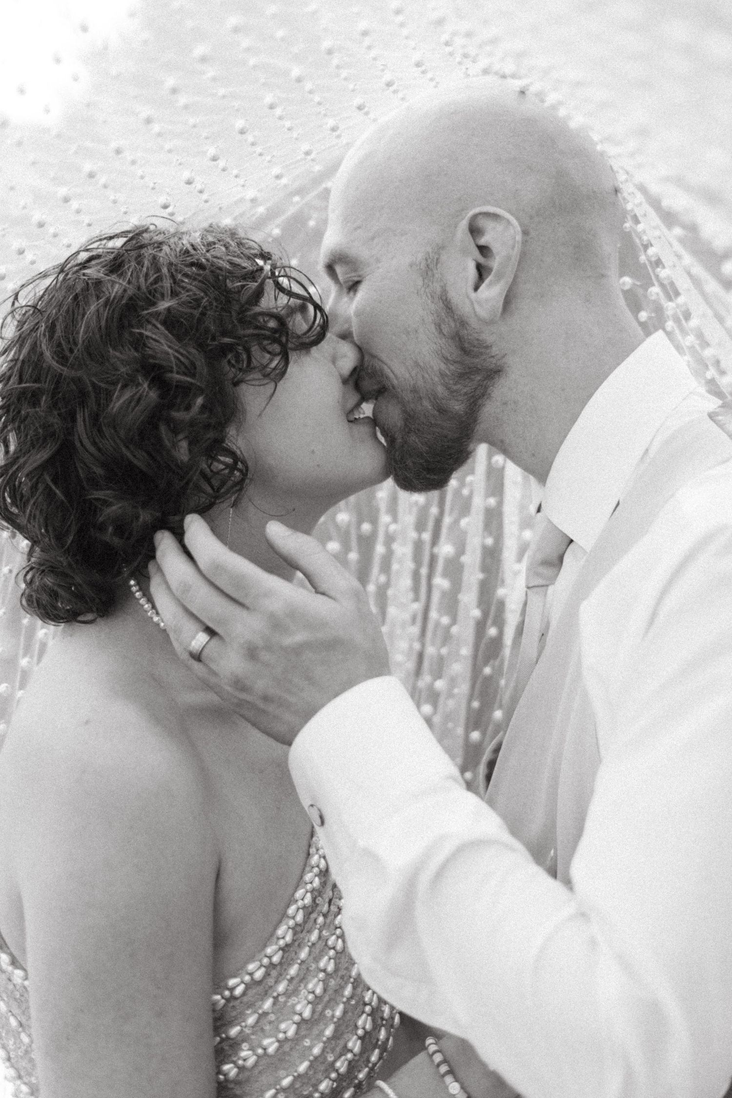 Black and white close-up of bride and groom sharing a tender kiss beneath a pearl-adorned wedding veil.
