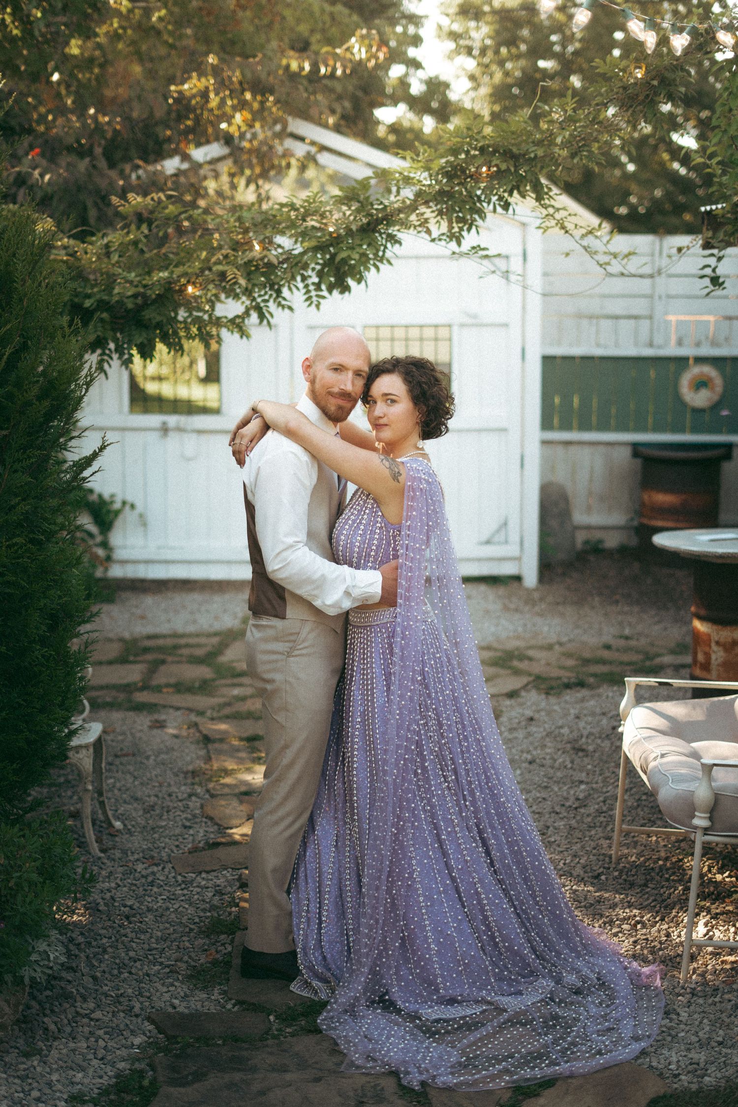 Bride in elegant lavender beaded gown embraces groom in white shirt near rustic white garden gates.