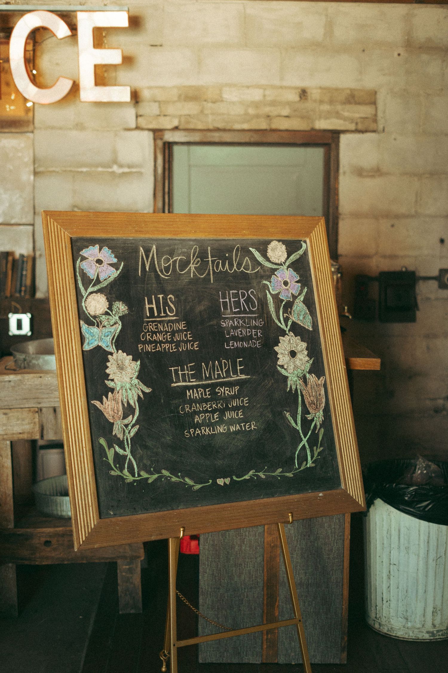 Chalkboard mocktail menu sign on easel listing his, hers, and the maple signature wedding drinks.