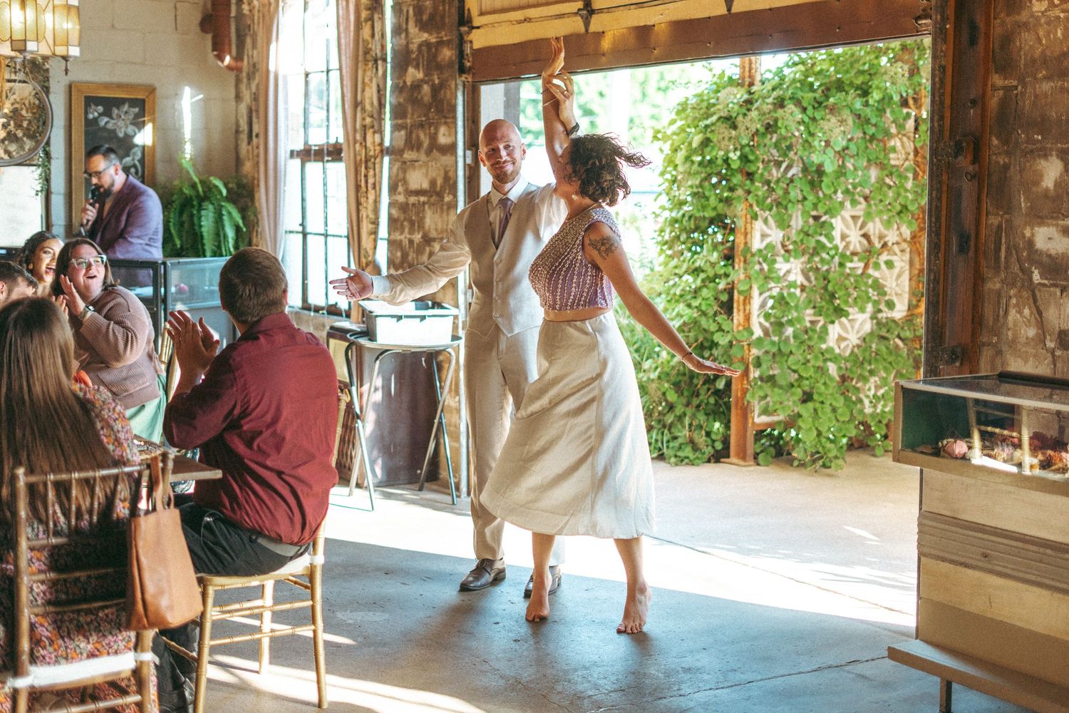 Bride and groom making a joyful grand entrance into rustic wedding reception as guests applaud.
