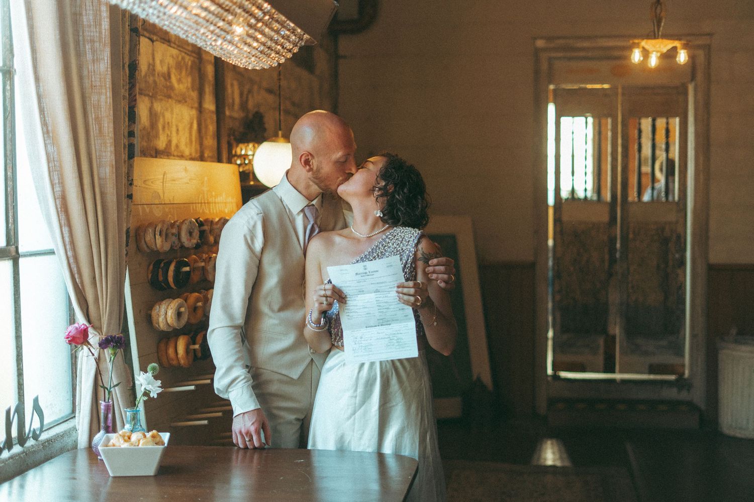 Couple sharing a kiss at a rustic wedding venue while holding their marriage certificate.