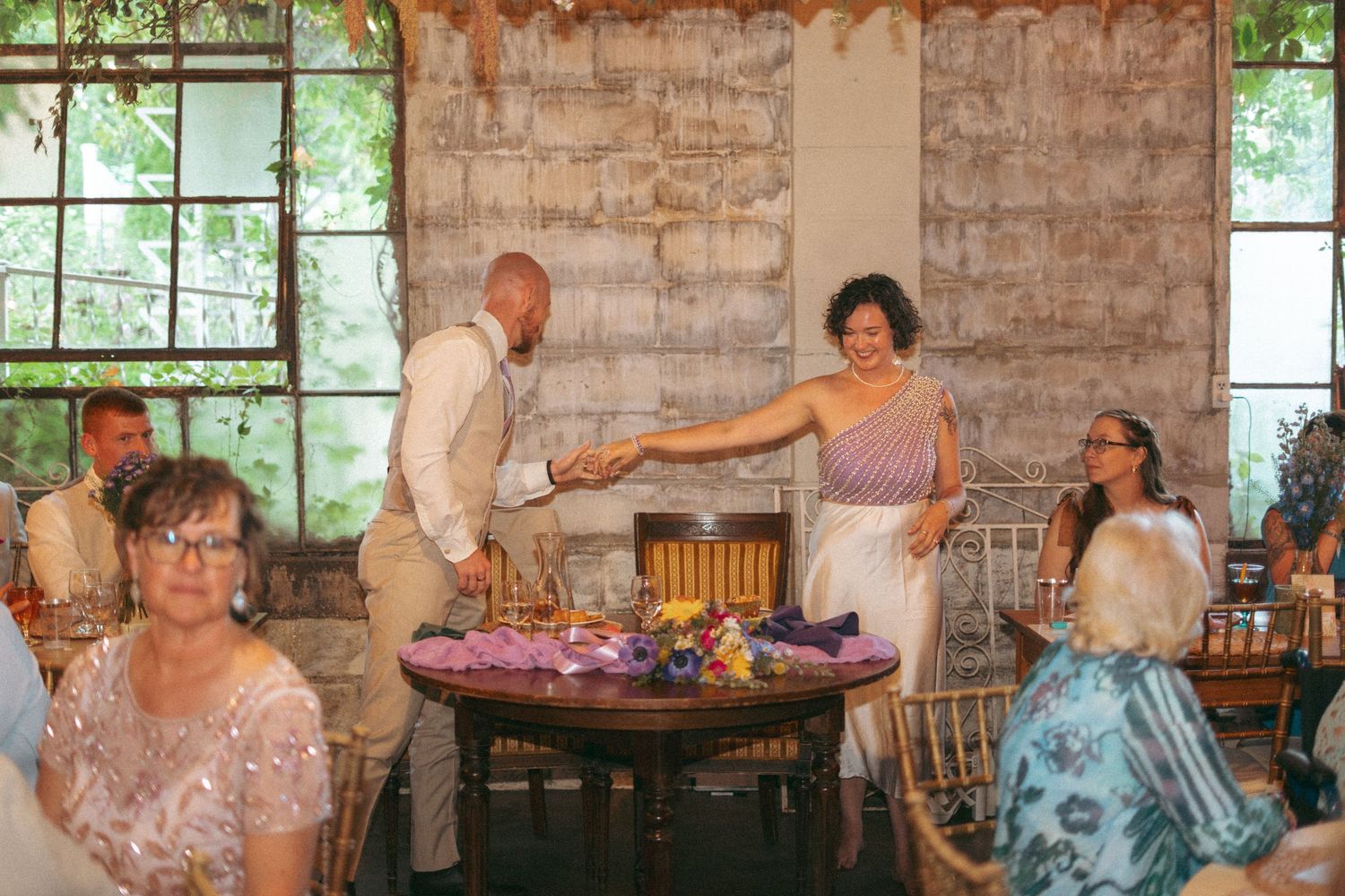 Newlyweds holding hands and dancing at their boho wedding reception with guests seated.