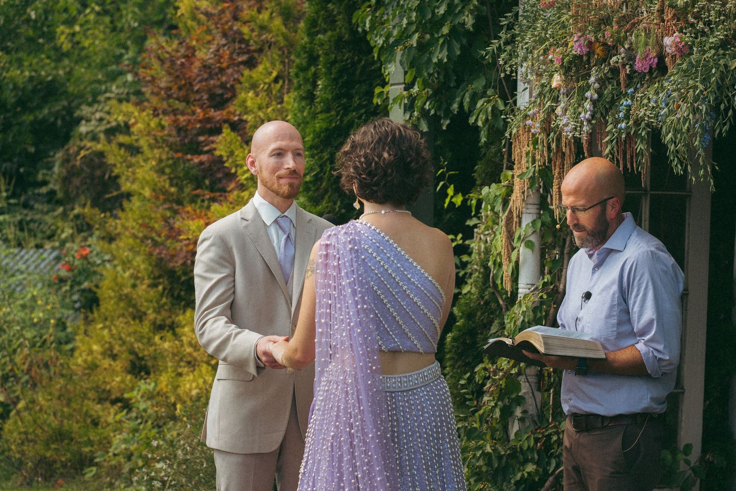 Groom gazes lovingly at bride in lavender dress while officiant reads at outdoor ceremony.
