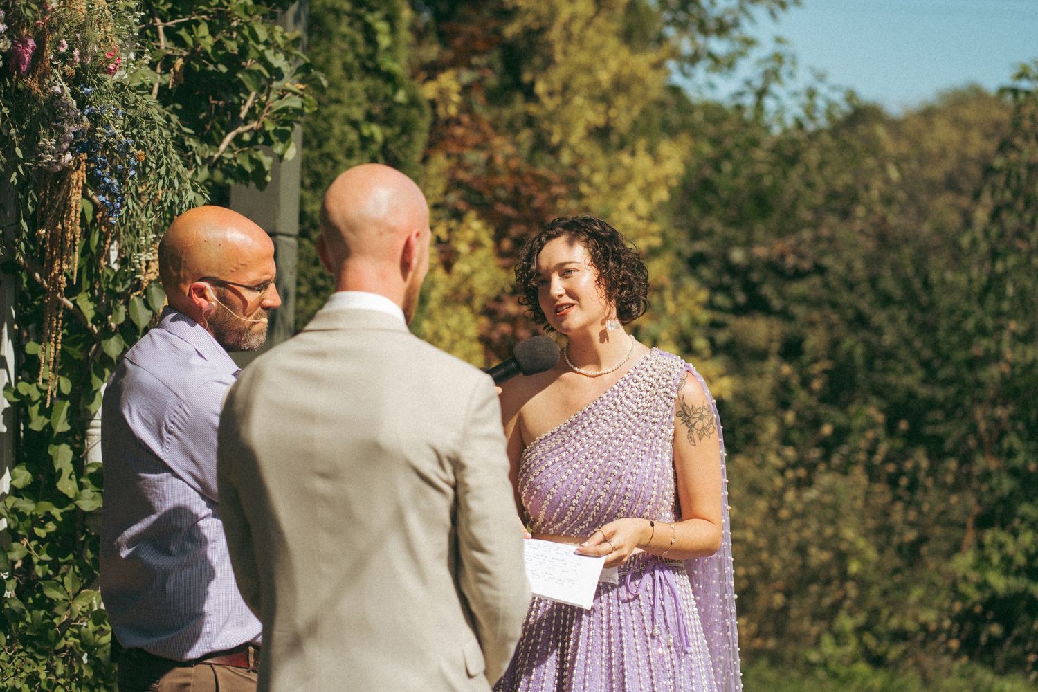 Bride in purple dress reads vows to groom at an outdoor garden wedding ceremony.