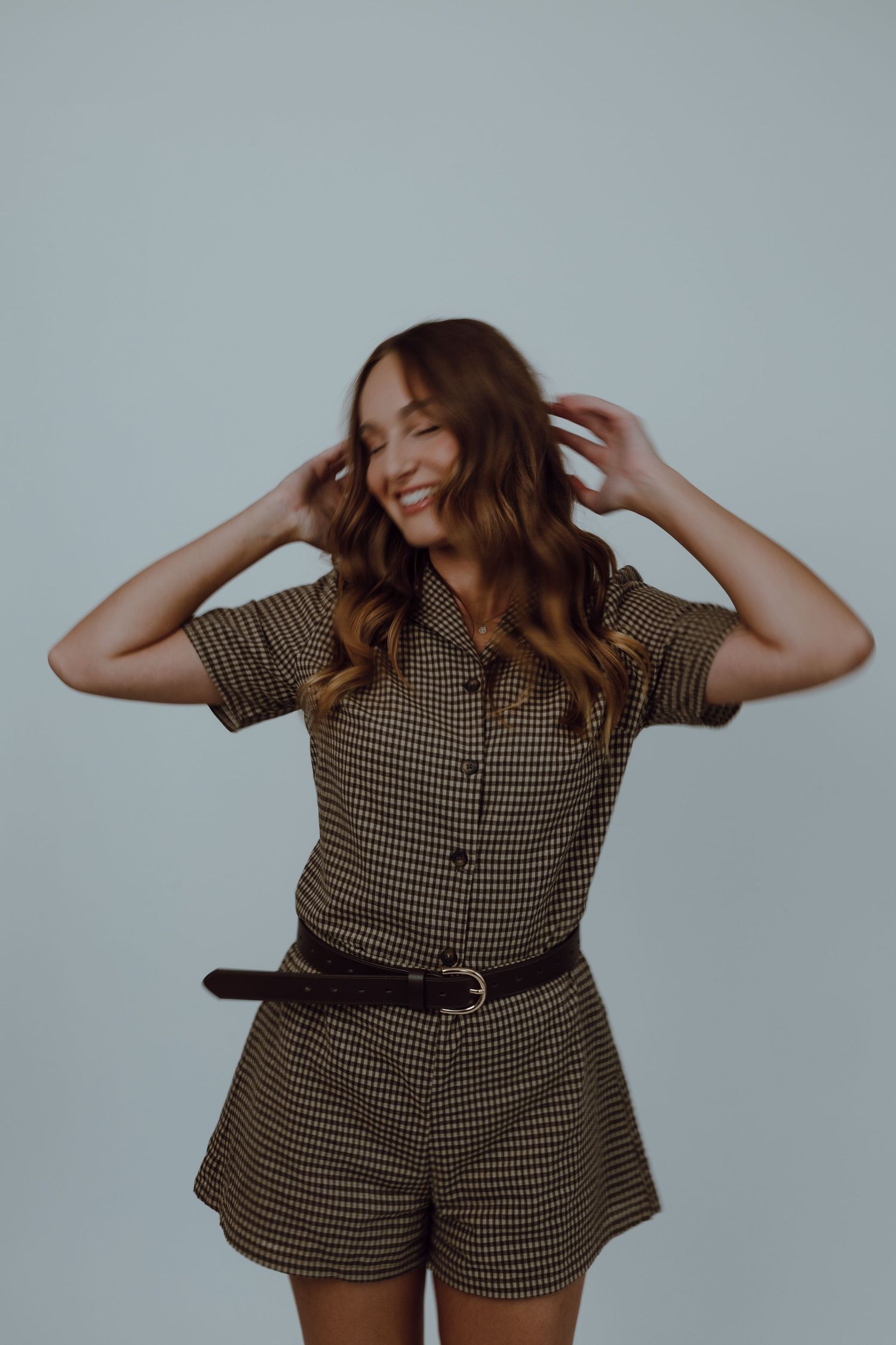 A person in a brown checkered romper poses playfully against a light gray background.