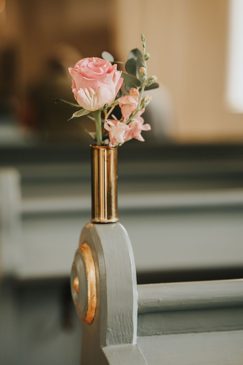 Pink roses elegantly arranged in a gold bullet casing vase create a striking contrast against a soft gray background.