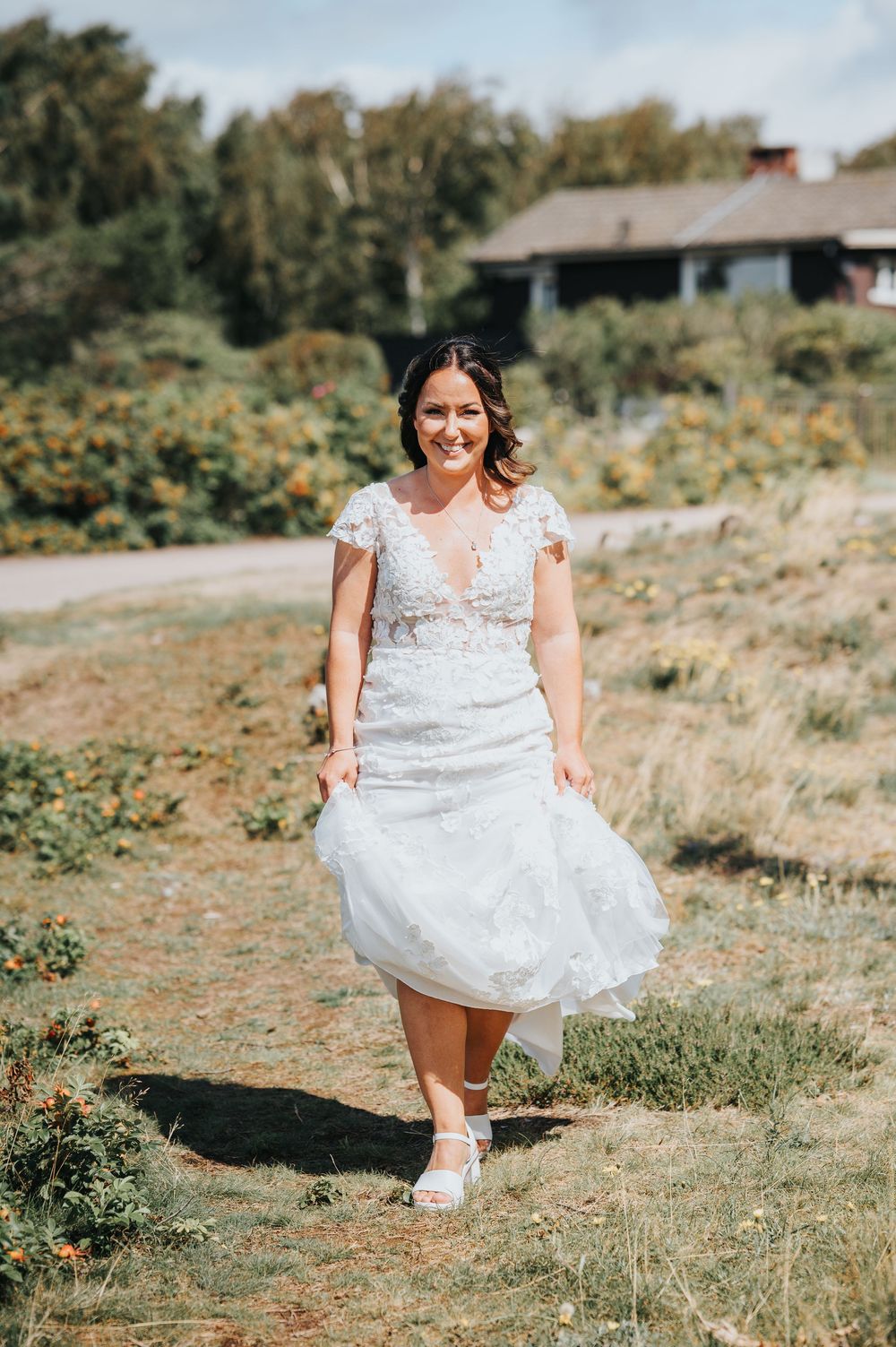 A person in a white lace wedding dress twirls playfully in a sunny outdoor field.
